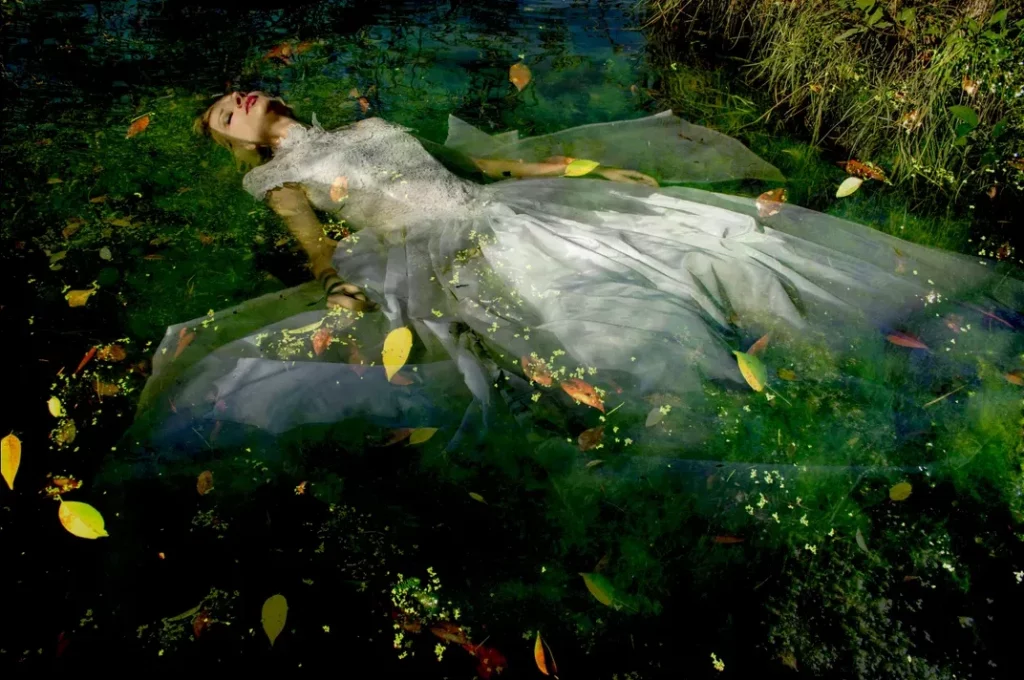 Glossy color photograph: Ophelia by Viet Ha Tran. A woman in white floats in a green river, surrounded by yellow leaves.