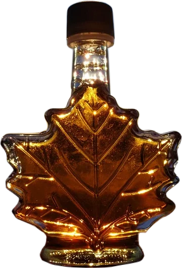 A glass bottle shaped like a maple leaf with a black cap filled with golden amber-colored maple syrup, backlit by sun.